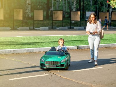 Mom controls children car with remote at walk with toddler child in public park. Toddler boy sits in green car holding steering wheel looking aside